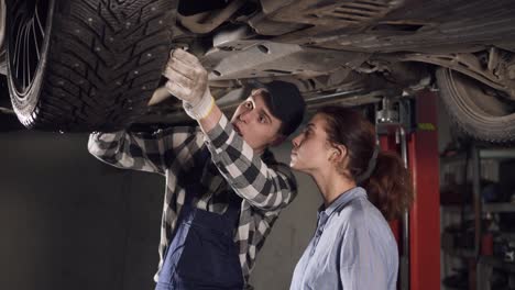 Handsome-mechanic-showing-his-female-client-all-the-problems-in-her-vehicle,-pointing-at-some-mechanisms-on-the-bottom-of-the-car.