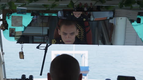 a view from inside a boat's control room, showing a reflection of a uniformed captain in the rearview mirror