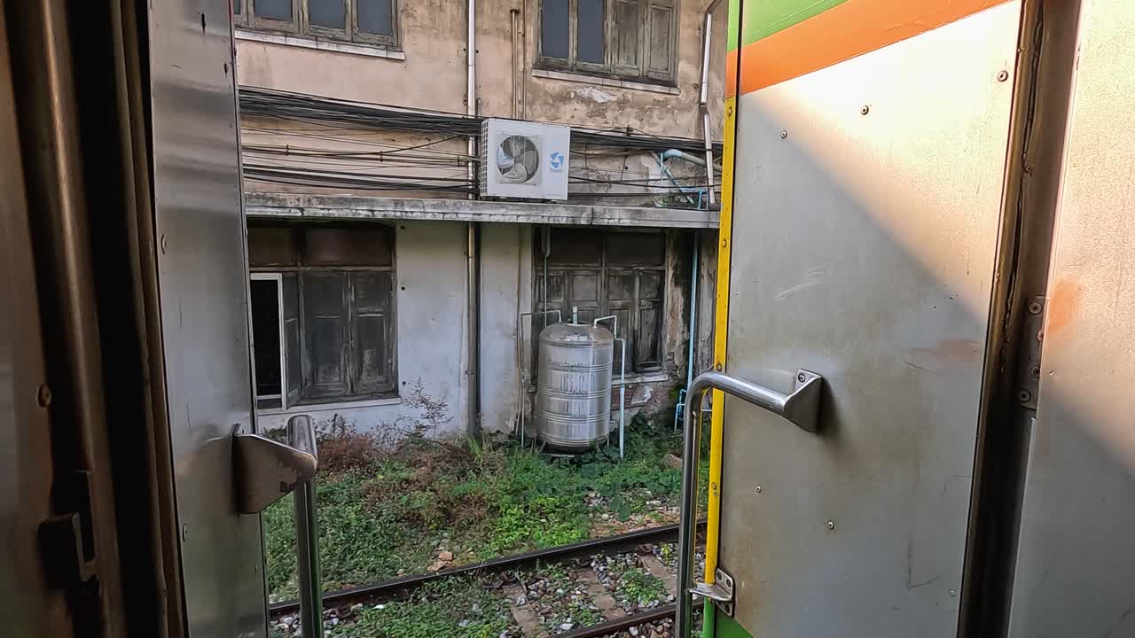 Passenger train door view of overgrown railway yard and weathered industrial buildings in Bangkok