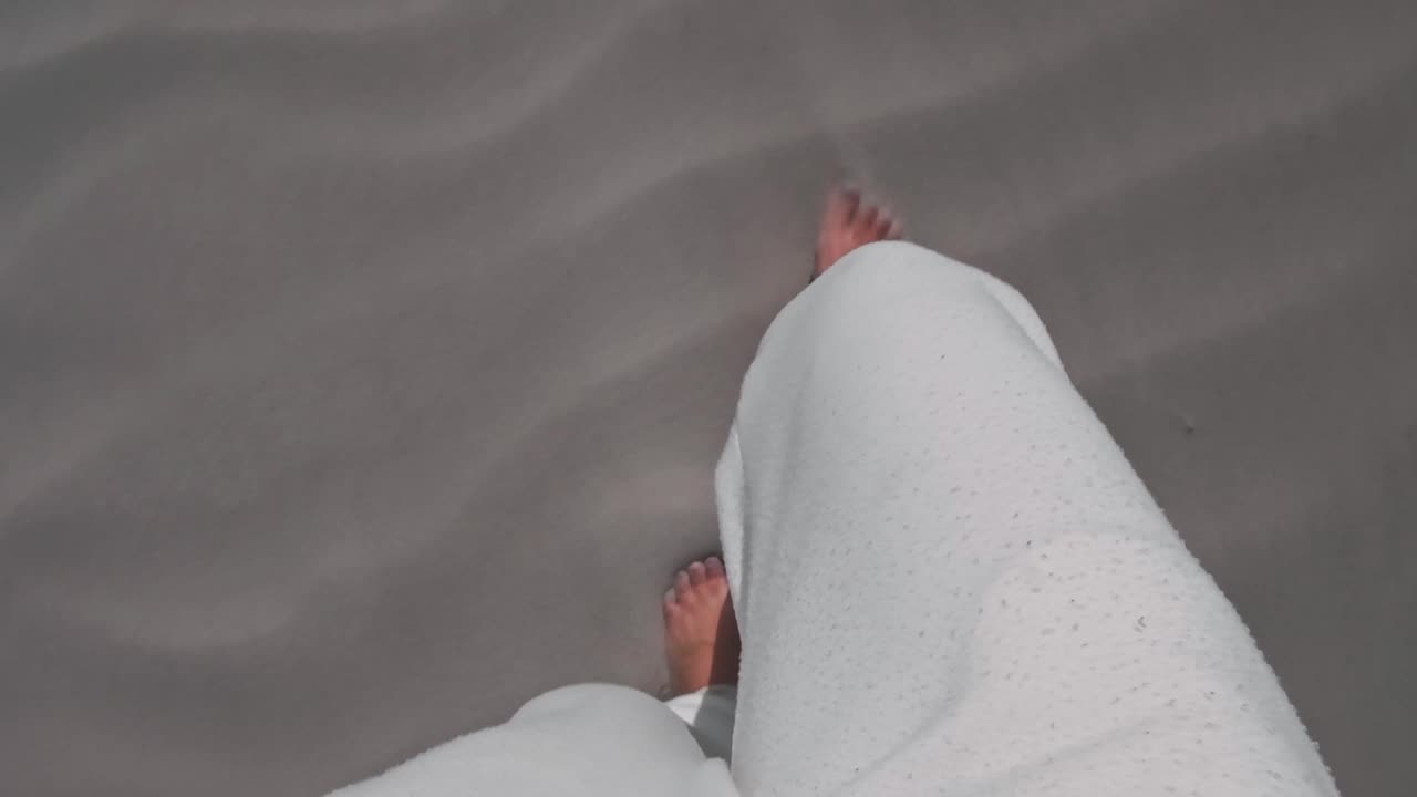 En person i hvide joggingbukser går barfodet på en hvid sandstrand