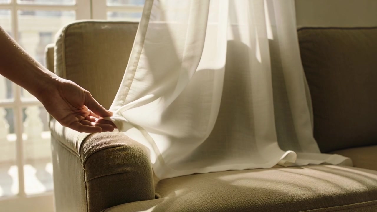 Entering bare hand smoothing sheer white curtain over beige armchair at window, creating even drape