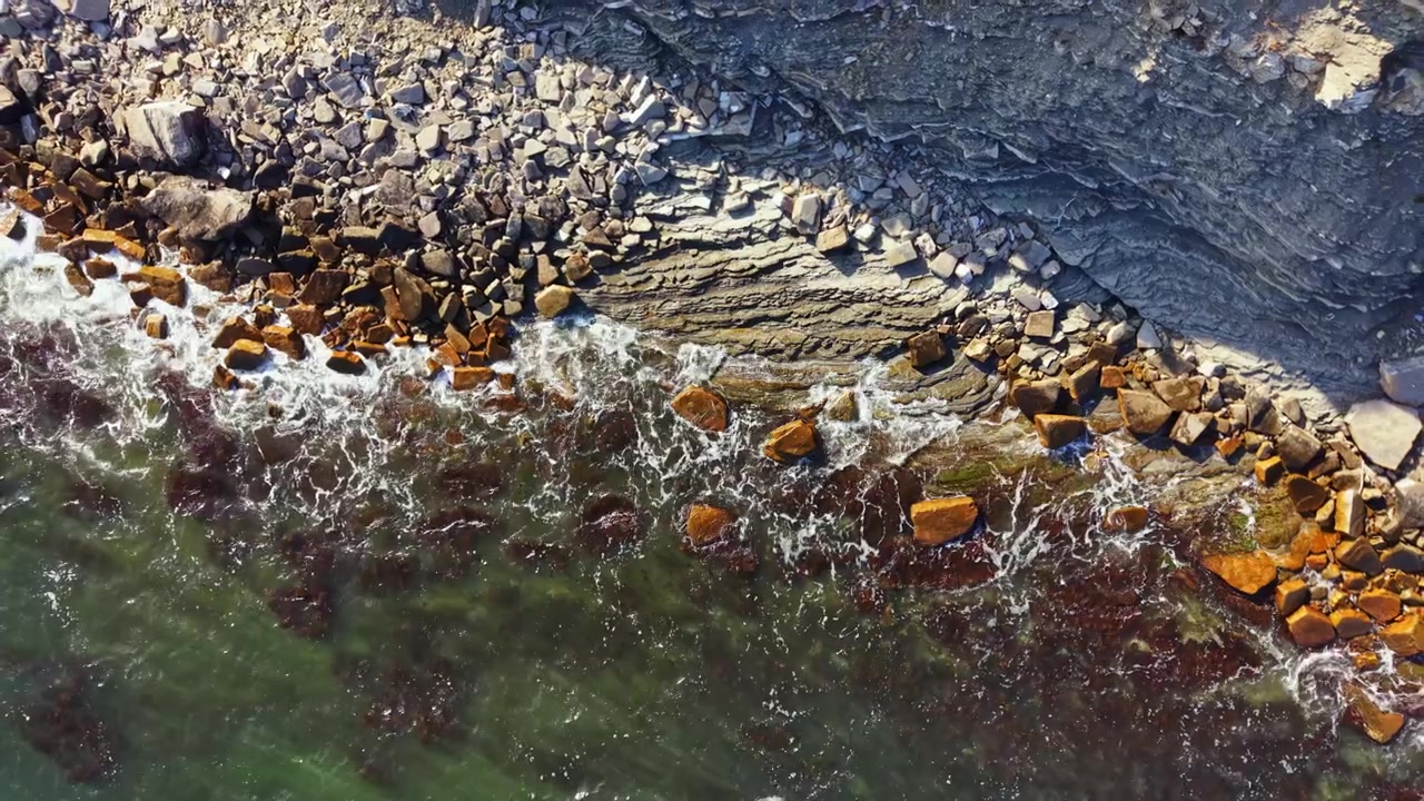 Waves crash against rocks along the shore in a coastal area during daylight hours near the ocean