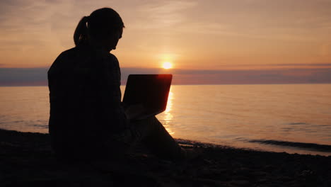 Silhouette-Einer-Frau,-Die-Bei-Sonnenuntergang-Mit-Einem-Laptop-Am-Meer-Arbeitet