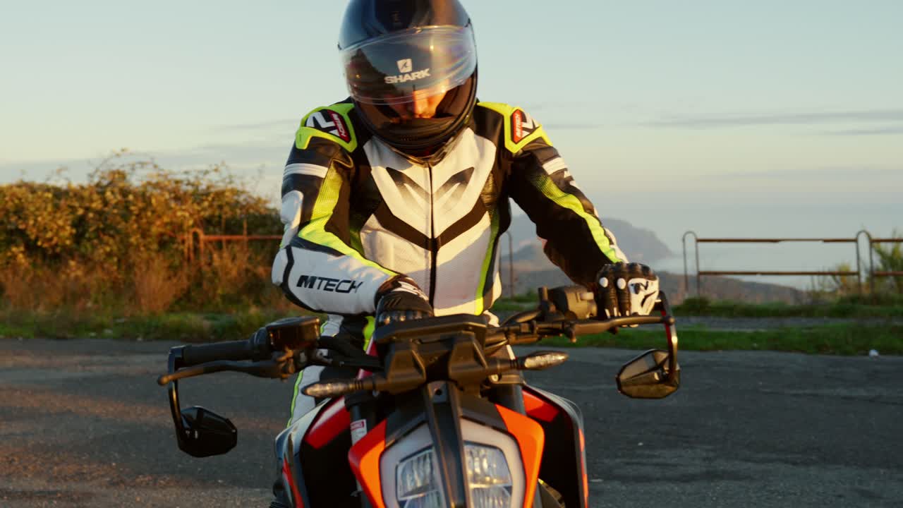 Motorcyclist adjusts his sport bike at sunset near a scenic coastal road, wearing full gear in slowmotion in Autumn