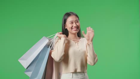 una mujer de compras asiática sosteniendo bolsas de compras antes de agitar las manos y sonreír a la cámara mientras está de pie frente a la pantalla verde