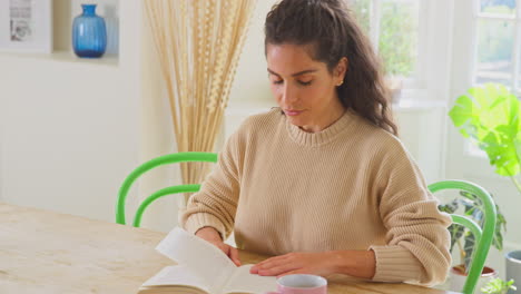 Frau-Entspannt-Sich-Zu-Hause,-Sitzt-Am-Tisch-Und-Liest-Ein-Buch-Mit-Heißem-Getränk