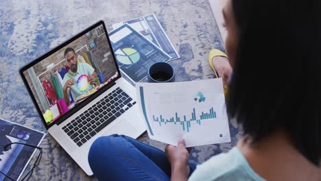 Mixed-race-businesswoman-sitting-on-floor-using-laptop-having-video-call-with-male-colleague