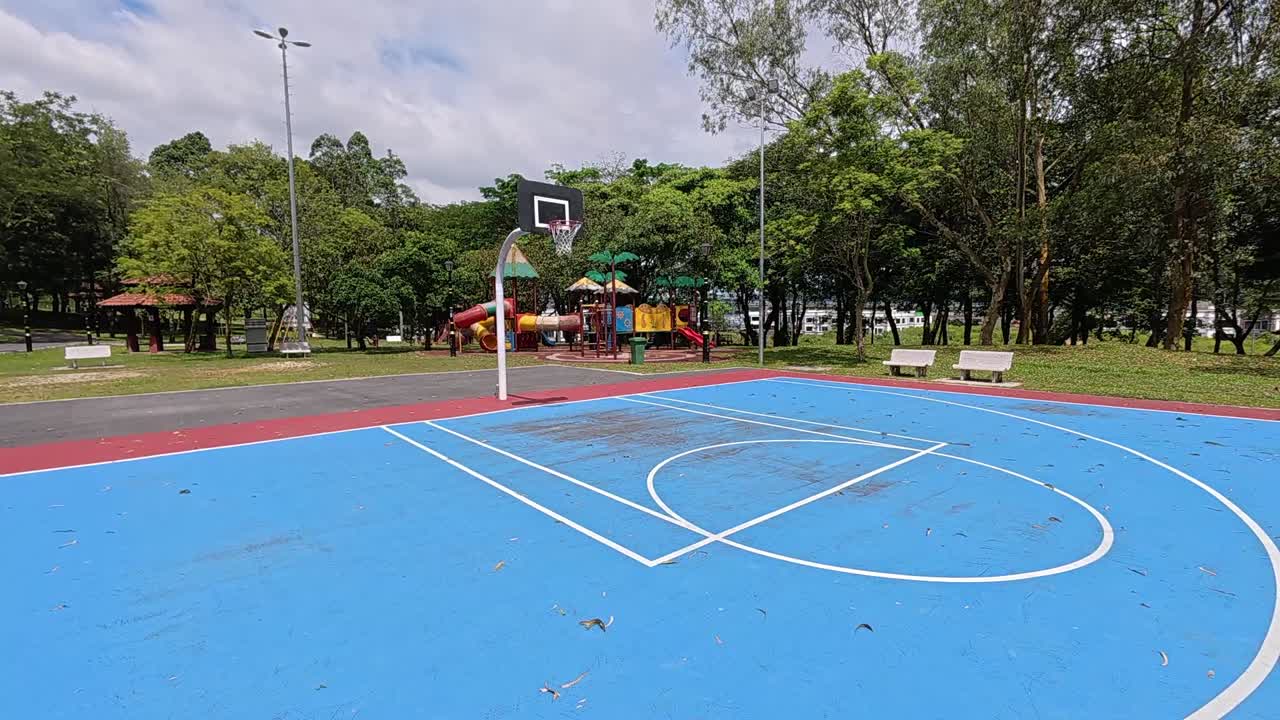 An empty basketball court in a tropical park, surrounded by lush green trees under a bright, sunny sky. Perfect weather.