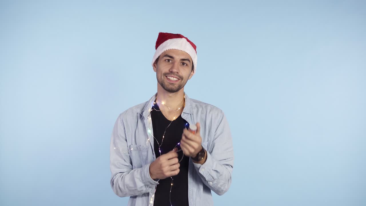 캐주얼 옷을 입고 머리에 산타 모자를 쓴 젊고 웃는 남자가 파란색 배경에 색종이 크래커를 폭발시키고 있습니다. 새해나 크리스마스를 축하하며 행복하게 지내세요