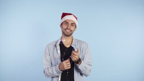 Hombre-Joven-Y-Sonriente-Con-Ropa-Informal-Y-Sombrero-De-Papá-Noel-En-La-Cabeza-Explotando-Una-Galleta-De-Confeti-Sobre-Un-Fondo-Azul.-Feliz-Celebración-De-Un-Nuevo-Año-O-Navidad