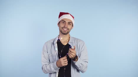 Young,-smiling-man-in-casual-clothes-and-santa-hat-on-a-head-exploding-confetti-cracker-on-a-blue-background.-Happy-celebrating-of-a-New-Year-or-Chrismas