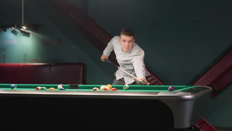 young man in white shirt and grey trousers leans over green pool table, gripping cue stick with intense focus as he strikes cue balls and walks. dark billiard room enhances concentration