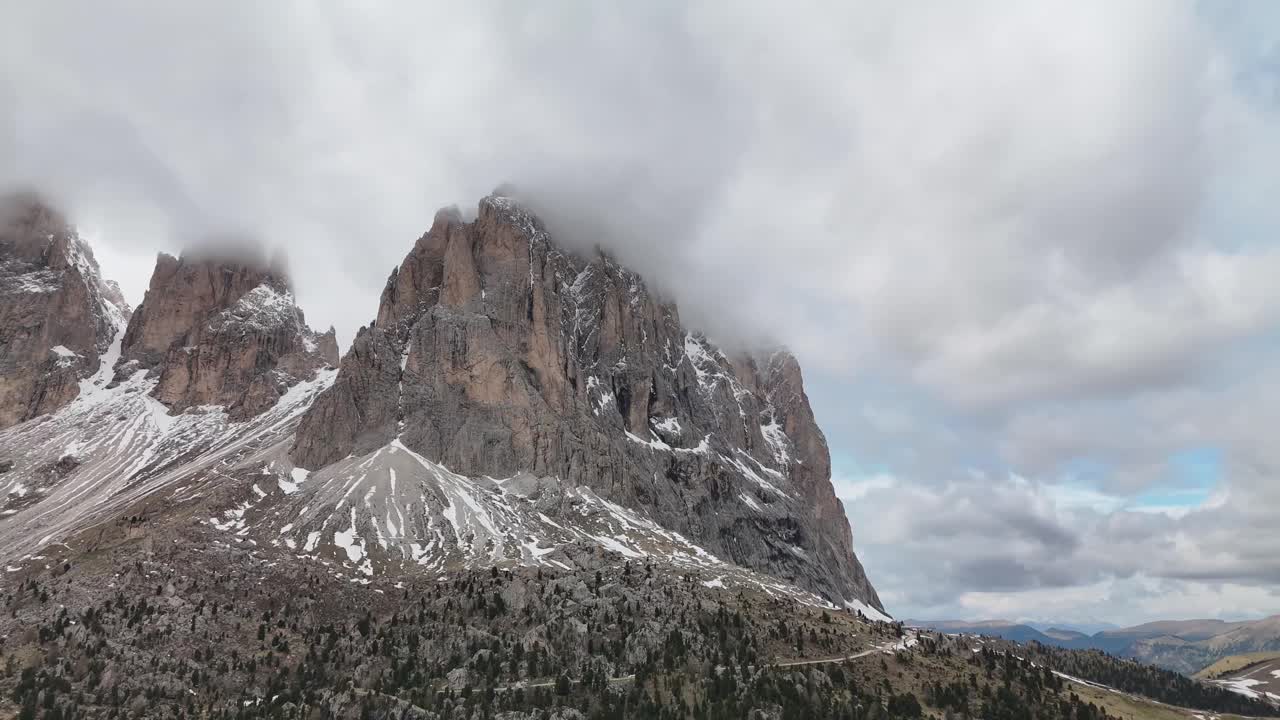 Drone aerial turn over Sassolungo Sella Pass in the Dolomites, Italy. Three towering peaks emerge through drifting clouds, creating a breathtaking alpine scene
