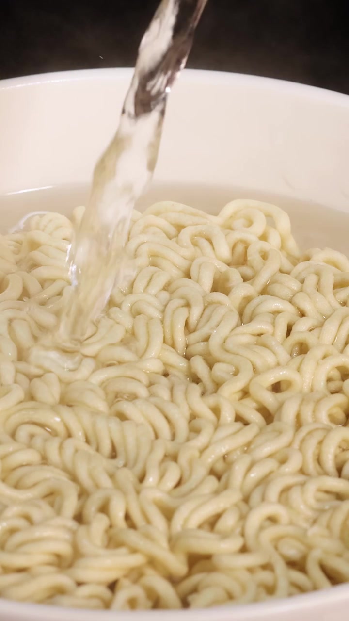 Un flusso costante di acqua calda viene versato su noodles istantanei secchi in una ciotola bianca, ripreso in primo piano con illuminazione brillante e uniforme e movimento minimo della telecamera.