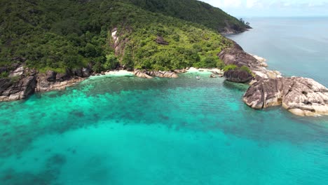 Drone-shot-heading-towards-sedcluded-rice-beaches-docked-boat-with-clients,-lush-national-park-and-turquoise-ocean,-Mahe-Seychelles-30-fps