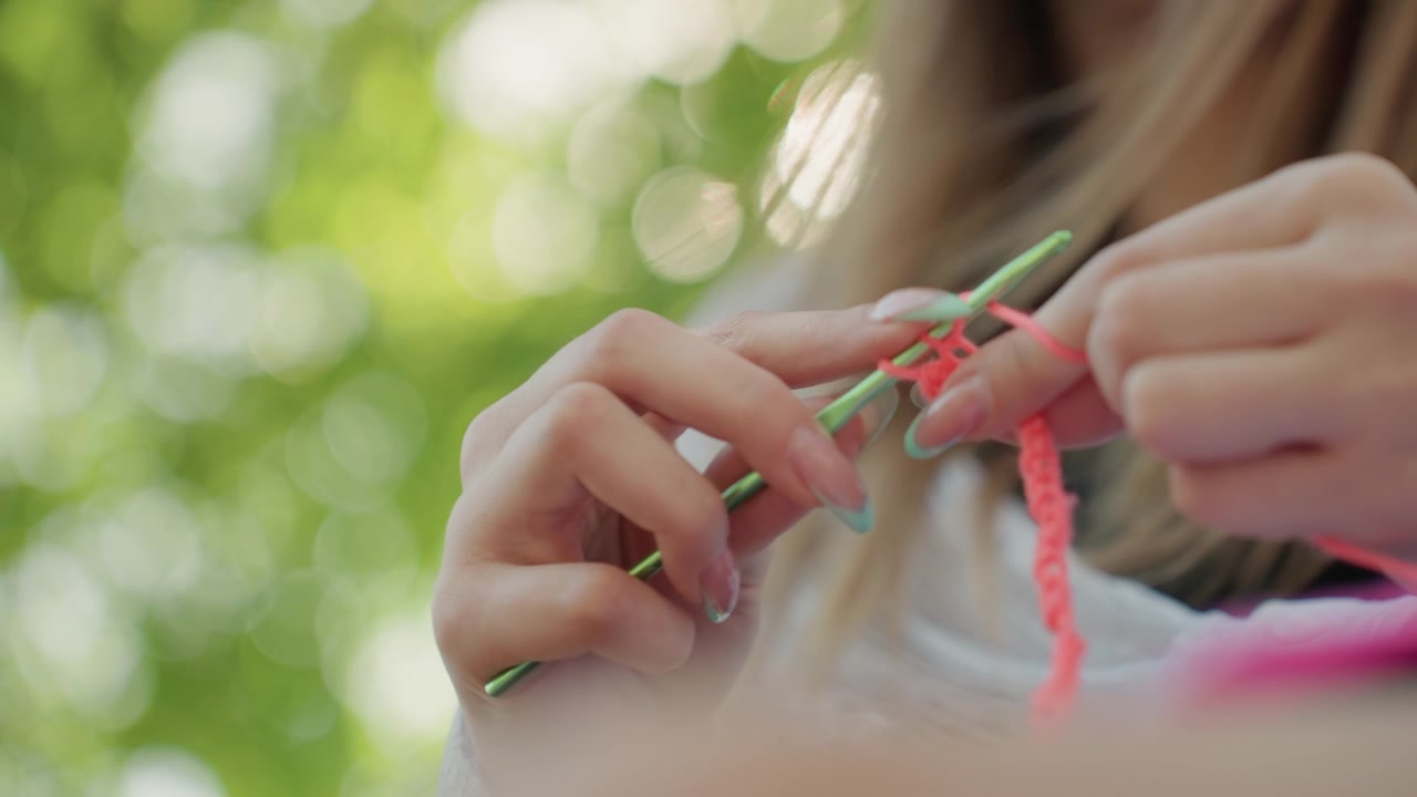Primo piano di giovane donna che lavora all'uncinetto con filato rosa e uncinetto verde, con le mani a fuoco contro uno sfondo esterno soffuso. La scena evidenzia i movimenti dettagliati delle mani mentre la donna crea motivi tessili.