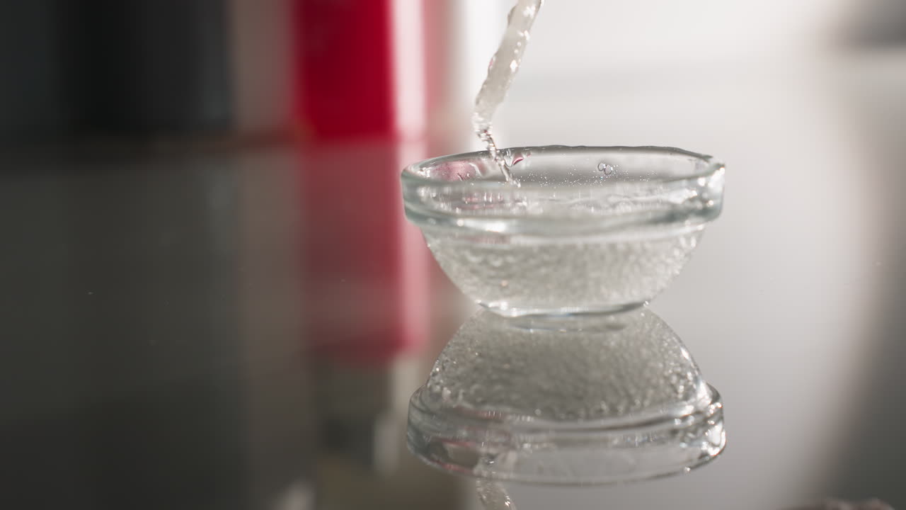 Face mask gel bubbles shimmer on clear surface as gloved hand of beauty technician stirs substance with silicone brush in small glass bowl creating reflective serene skincare prep scene