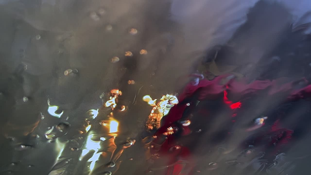 primer plano del desempañado del parabrisas con calefacción debido al mal tiempo con lluvia y aguanieve deslizándose hacia abajo en la ventana empañada y luces borrosas del tráfico de automóviles en segundo plano