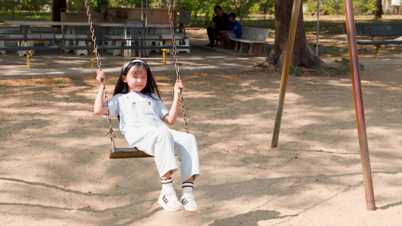 Bambino asiatico in salopette di jeans si dondola felicemente in un parco giochi all'aperto nel Parco Lumpini, Bangkok.