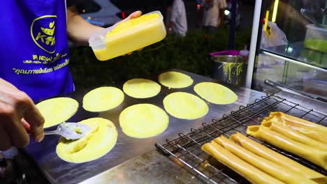 A vendor skillfully prepares crispy pancakes on a street stall in Bangkok, using a batter dispenser under bright lighting