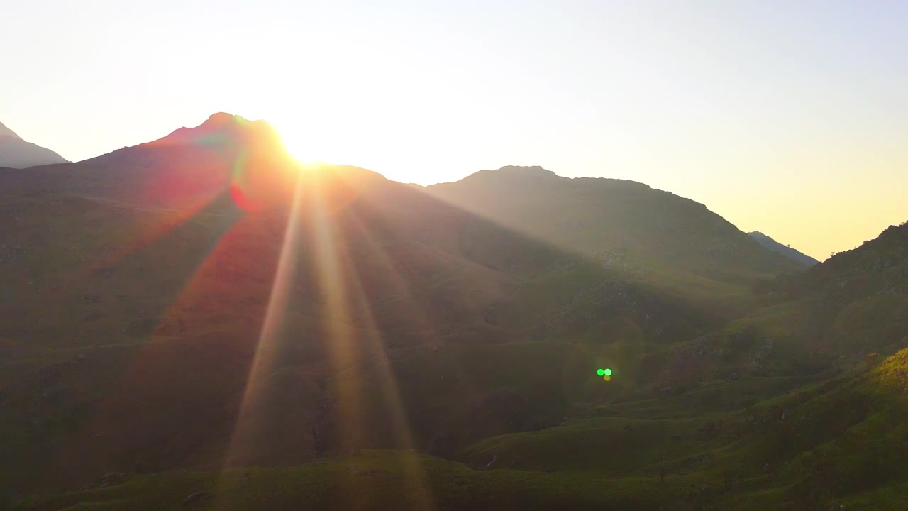 Drone tracks laterally as the setting sun emerges behind a mountain ridge, casting warm golden rays across the green slopes of Mulanje in Malawi.