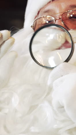 santa claus with a magnifying glass. elderly santa with white beard looking into glass in his residence. close-up. christmas concept. vertical video