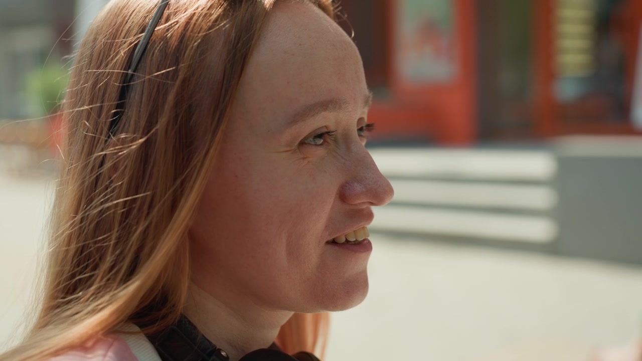 Woman with long hair wears headphones and pink shirt walking outdoors in daylight. background shows urban elements like buildings or storefronts suggesting everyday street scene. Soft
