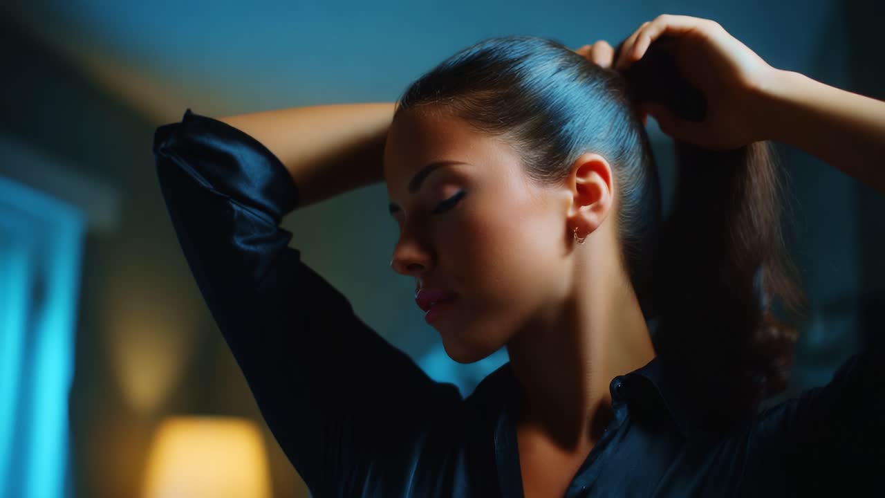 A close-up view of a young woman gracefully pulling her hair into a ponytail in a softly lit room, showcasing her profile and the elegance of her movements while dressed in a black shirt, capturing a moment of poised beauty