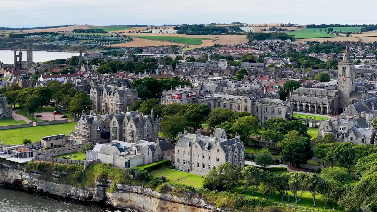 Drone glides over medieval ruins, university buildings, coastline, and town under soft daylight