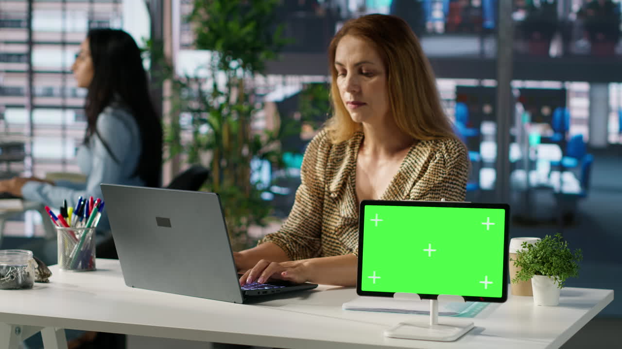 Woman working on laptop with tablet green screen in office