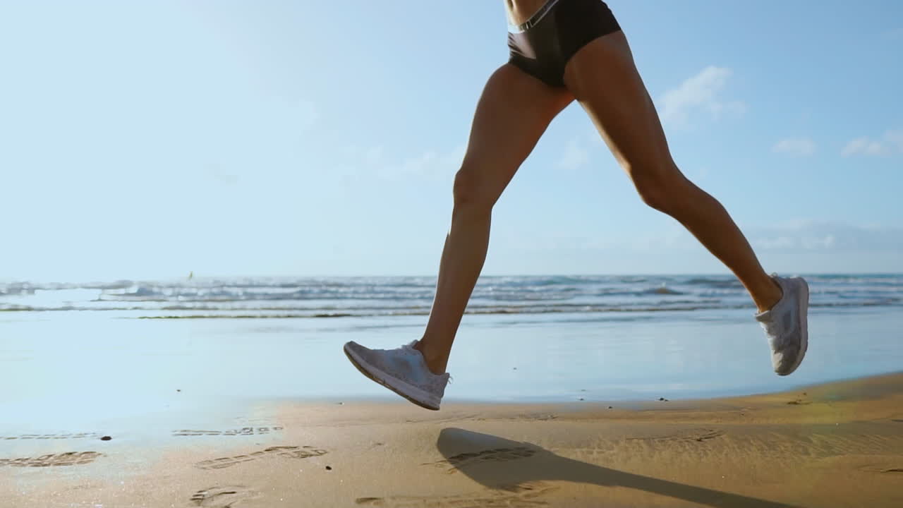 Close-up of leg sporty woman in white sneakers running along beautiful sandy beach, healthy lifestyle. in slow motion