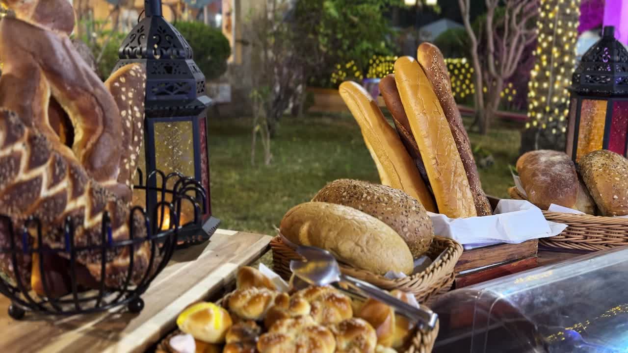 Baguette, pagnotte e panini assortiti su un tavolo da buffet rustico con il bagliore delle lanterne e le luci del giardino di notte, angolo pane per catering all'aperto