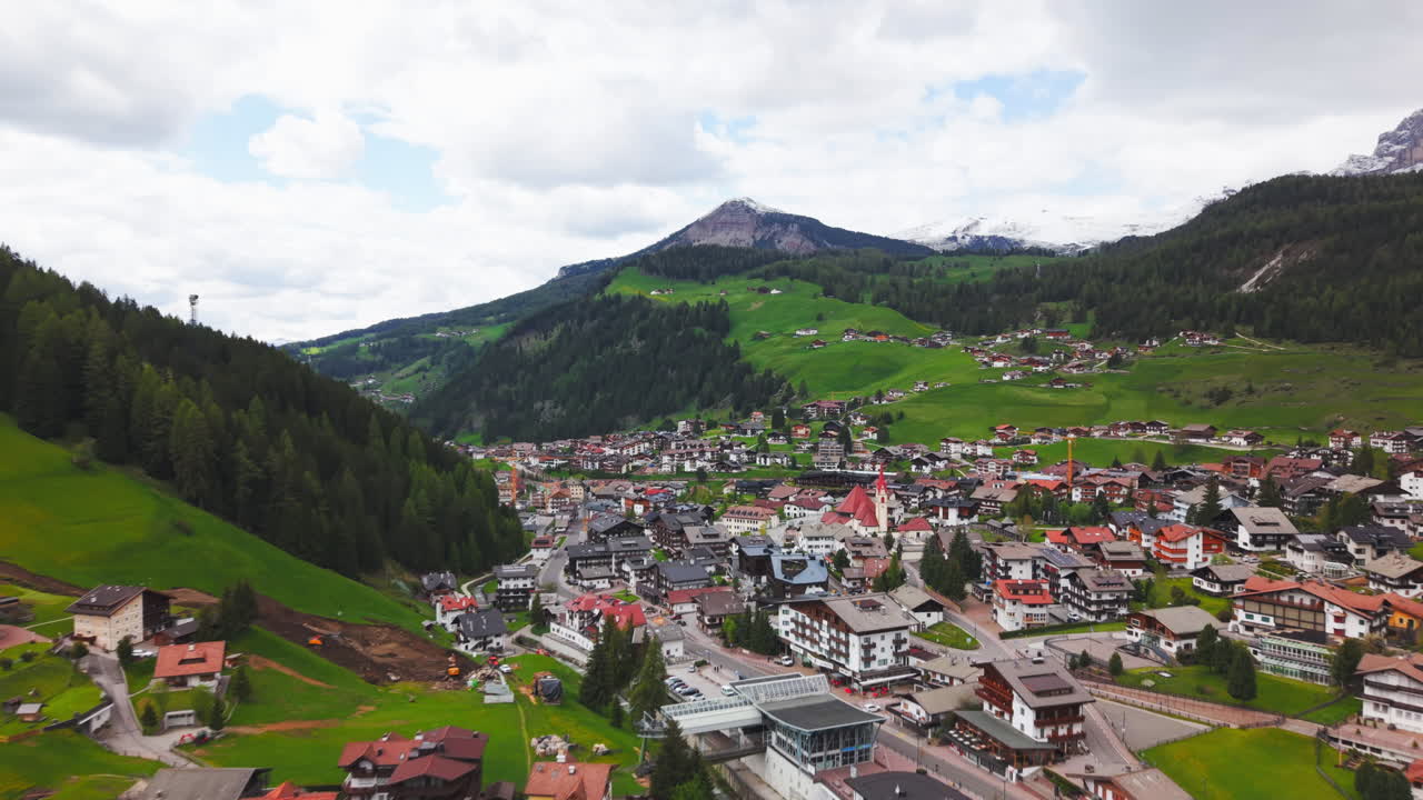 Drone footage of a picturesque alpine village surrounded by green hills, forested mountains, and snow-capped peaks in the Dolomites, Italy