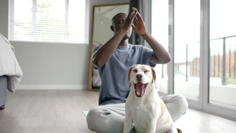 african american man doing yoga and meditating, with his pet dog at home, slow motion