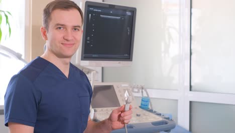 a man in blue scrub attire is gesturing with a syringe near an ultrasound machine in a room.