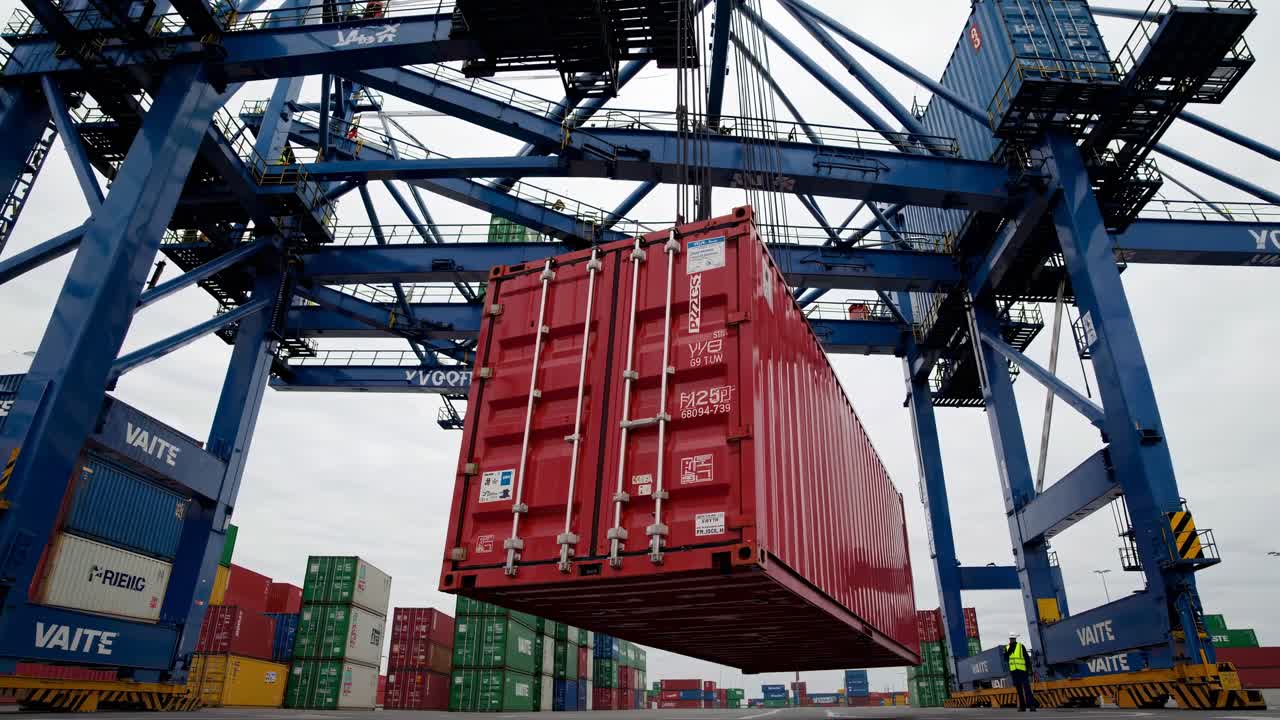 Low-angle shot of a red shipping container being lifted by a crane in a busy port