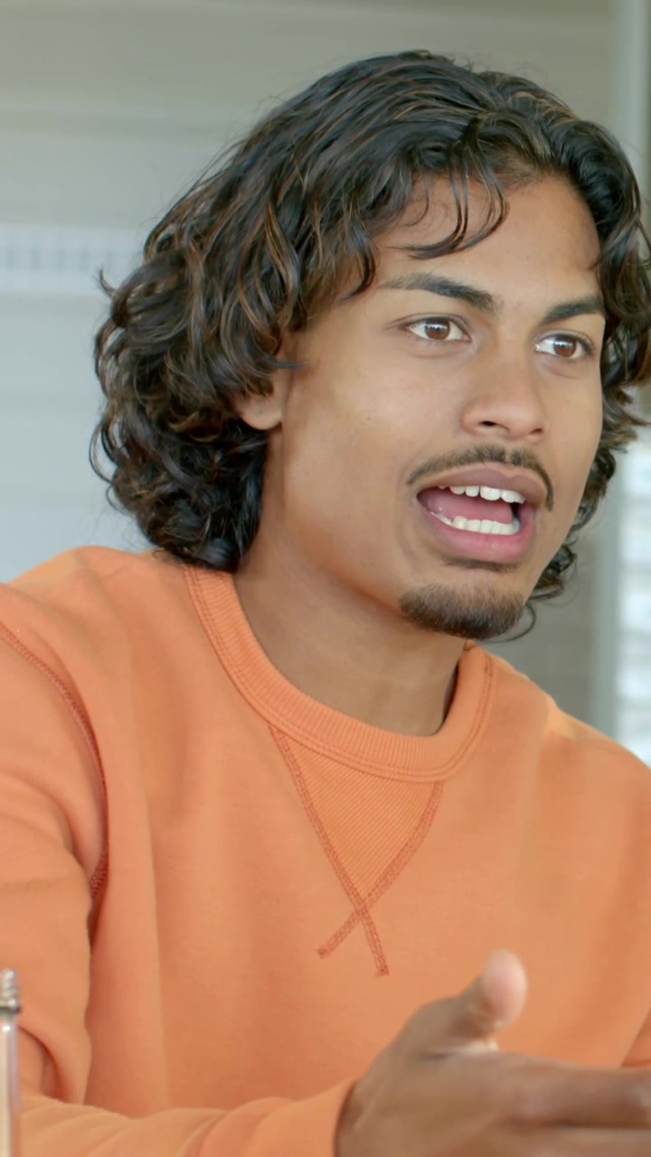 Vertical video: Turning man in orange talking, gesturing at home after offscreen prompt with bottle