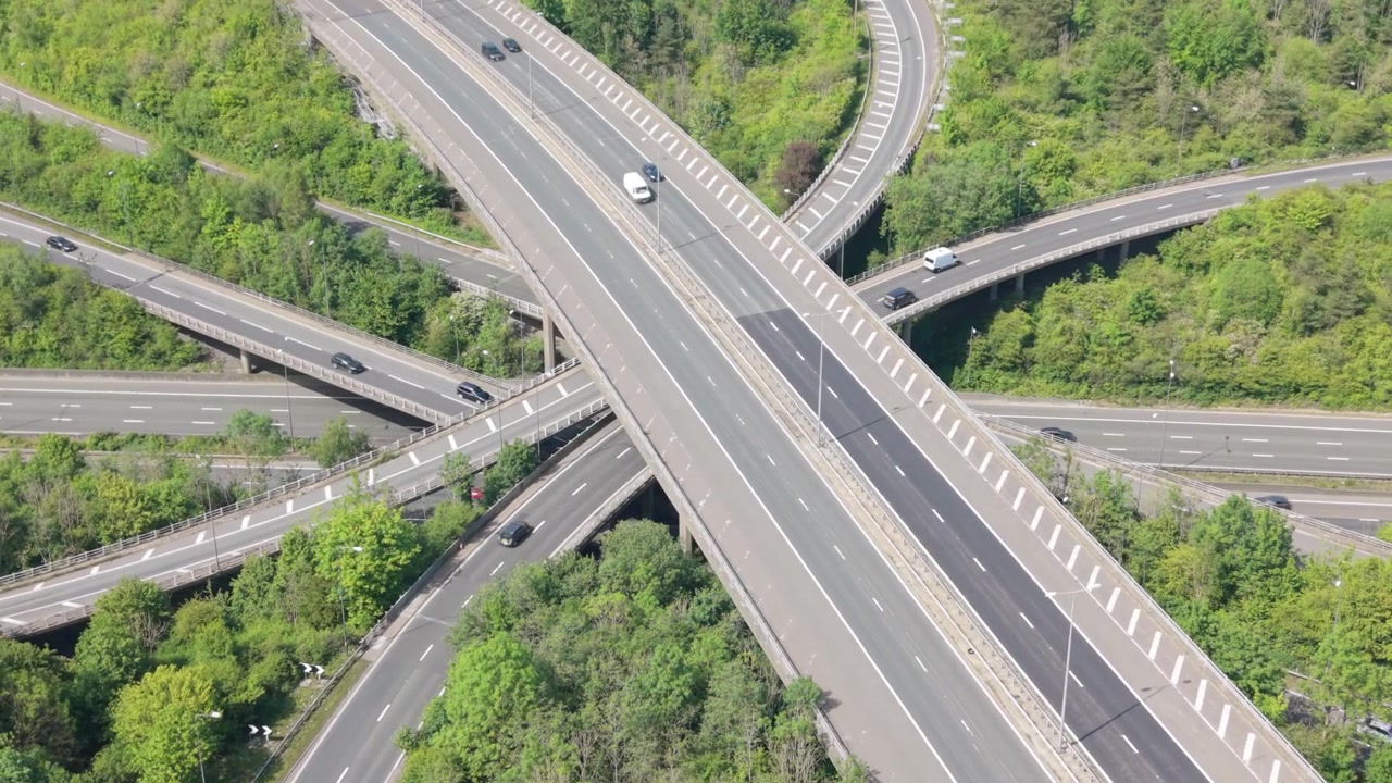 Aerial perspective of a complex multi-level highway interchange. Numerous cars travel across interconnected roads, surrounded by abundant green foliage during bright daylight.