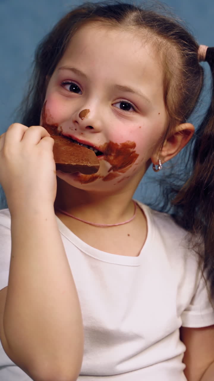 Little girl with dirty face eats bar of chocolate. Funny child with two ponytails is biting tasty black chocolate on blue studio background. Vertical video