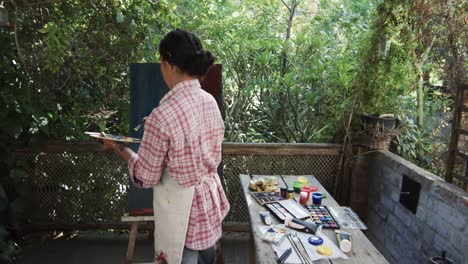 biracial female artist in apron painting on canvas in sunny garden, slow motion