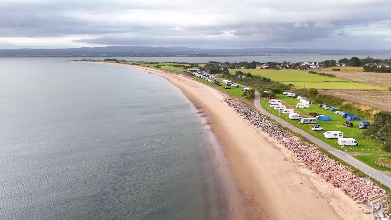 Droneoptagelser glider over en sandet kystlinje, roligt hav og en campingplads under overskyet himmel i Rosemarkie, Highland, Skotland. Jævn sideværts kamerabevægelse.