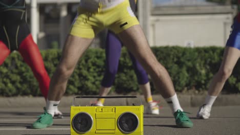 unrecognizable group of fit caucasian man and women jumping at the background to disco music from yellow vintage tape recorder standing on road at front. 1980s sportive lifestyle concept.