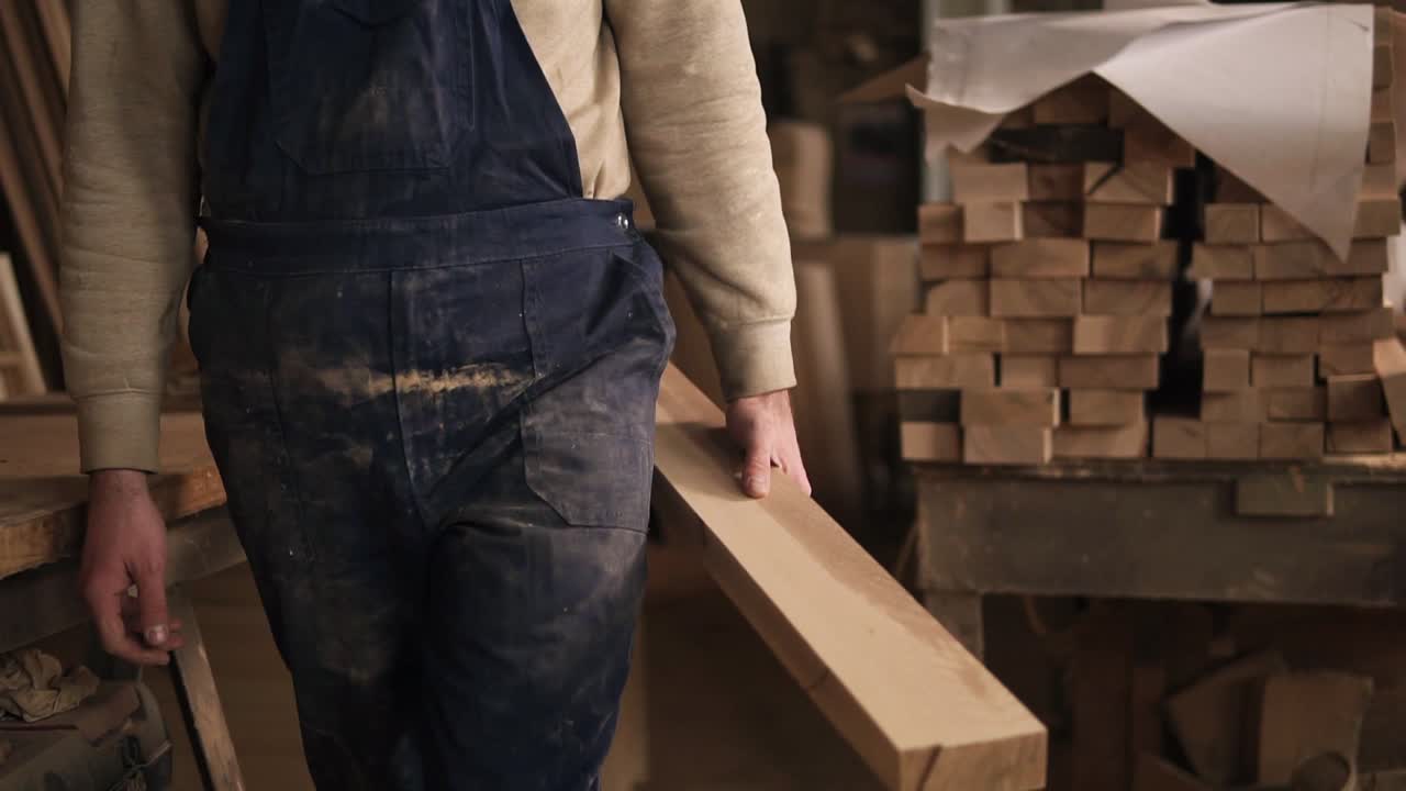 el carpintero en el taller va a la máquina de rectificar con una barra de madera. un hombre en mono azul. su polvoriento de trabajar con una madera . sin cara