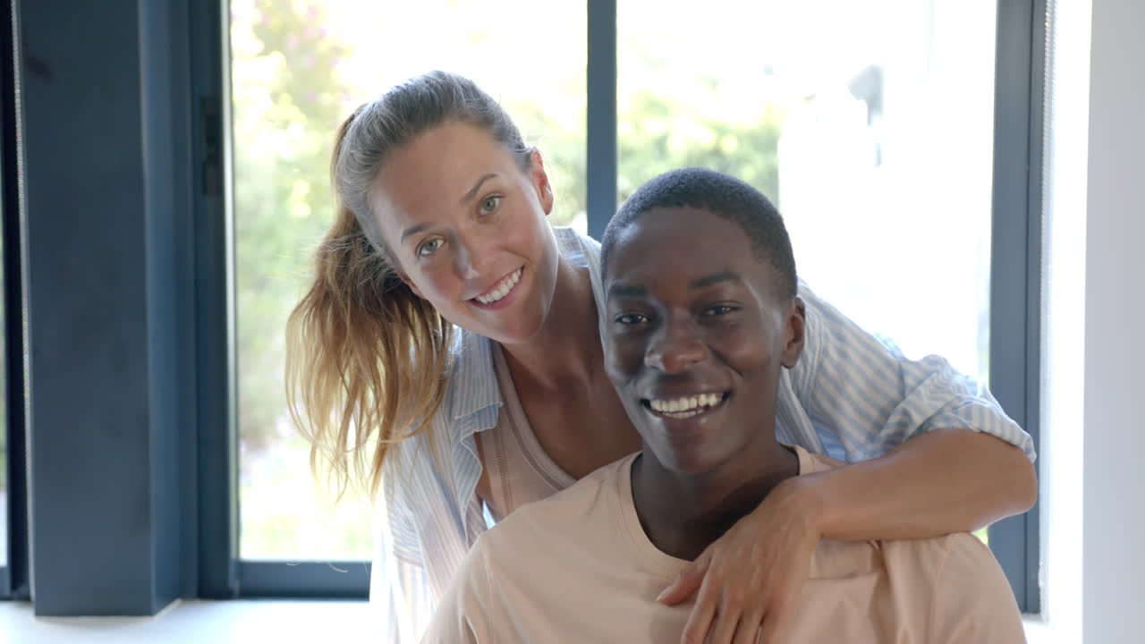 Coppia multietnica che si abbraccia e sorride vicino alla finestra, a casa