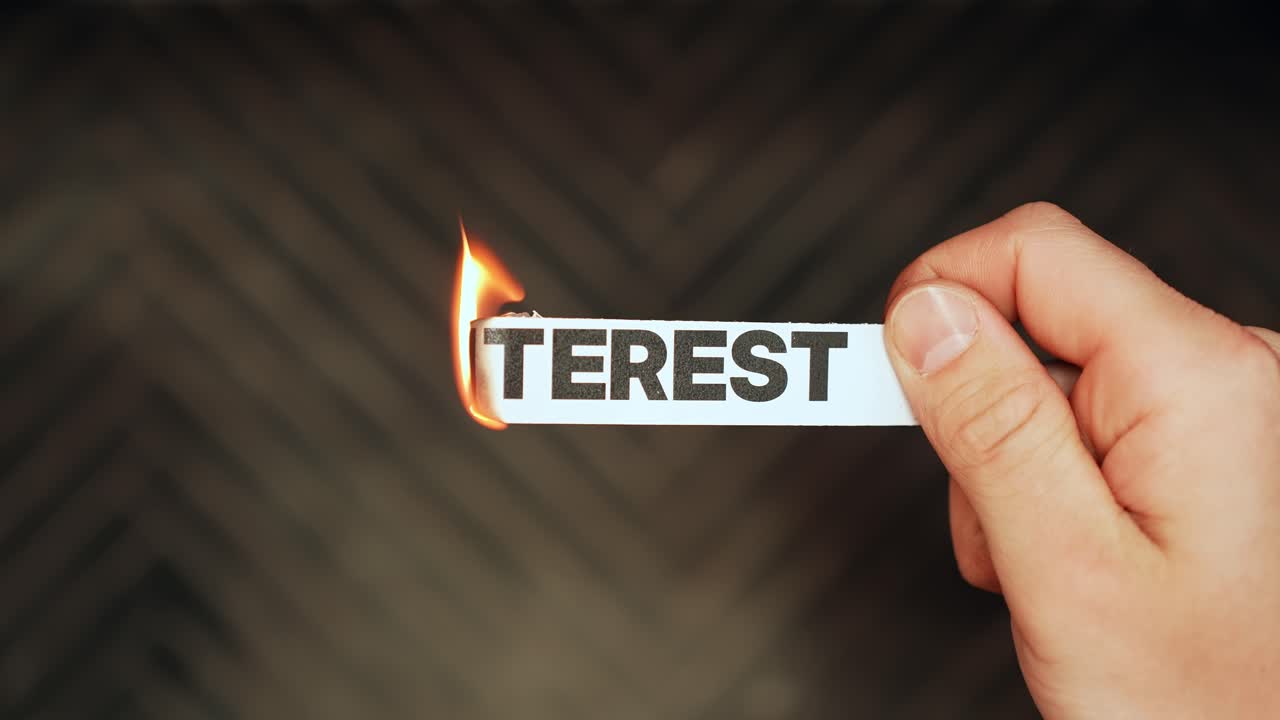 Hand holds a paper label reading “INTEREST” as flame burns the edge, symbolizing rising borrowing costs, debt pressure, and financial stress. Vertical close-up