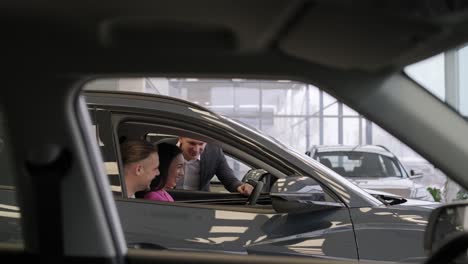 young car salesman showing to young couple new automobile at dealership salon.