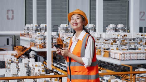 vista lateral de una ingeniera asiática con casco de seguridad usando un teléfono inteligente y mirando a su alrededor mientras está de pie en el almacén con estantes llenos de productos de entrega