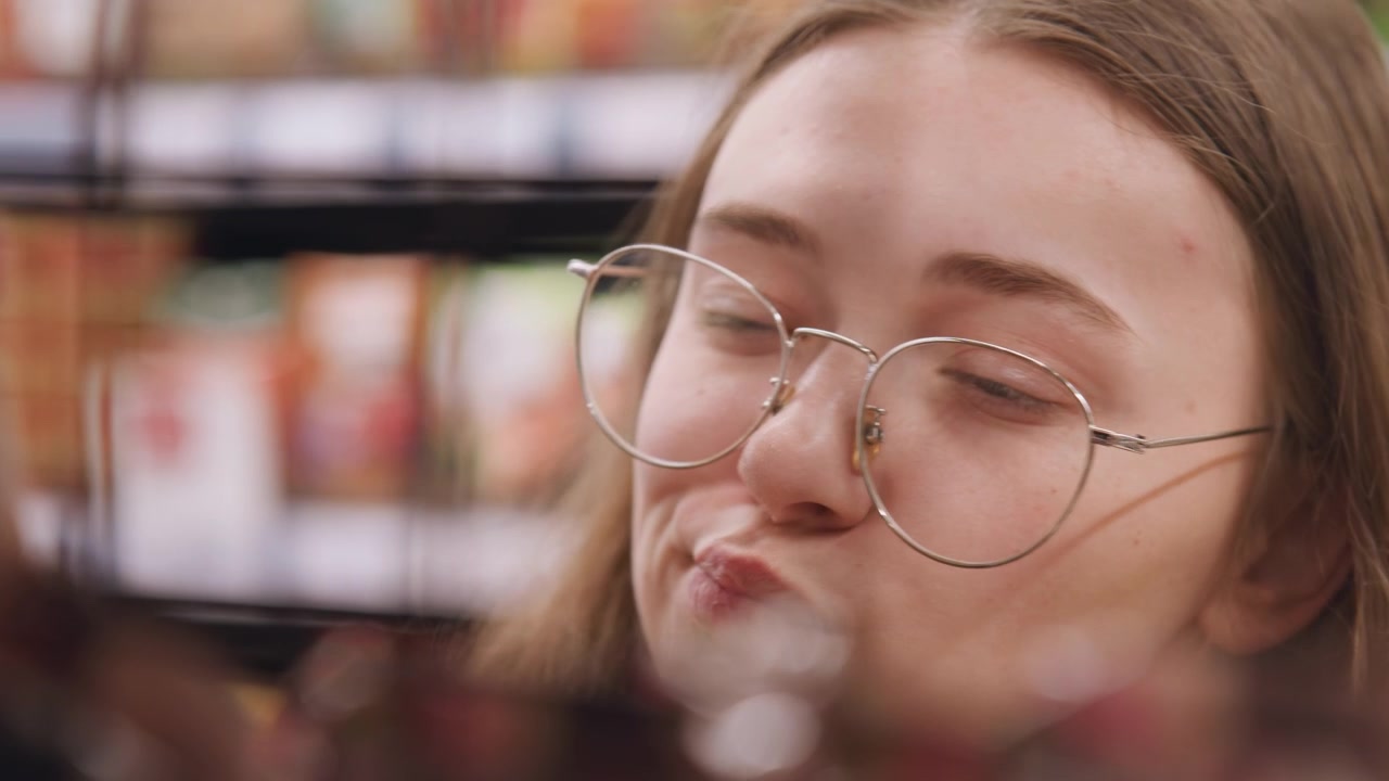 White woman comparing product choices on shelf with round glasses and steady gaze label reader checking nutrition, budget shopper weighing price, curious consumer reaching for snack jar while studying