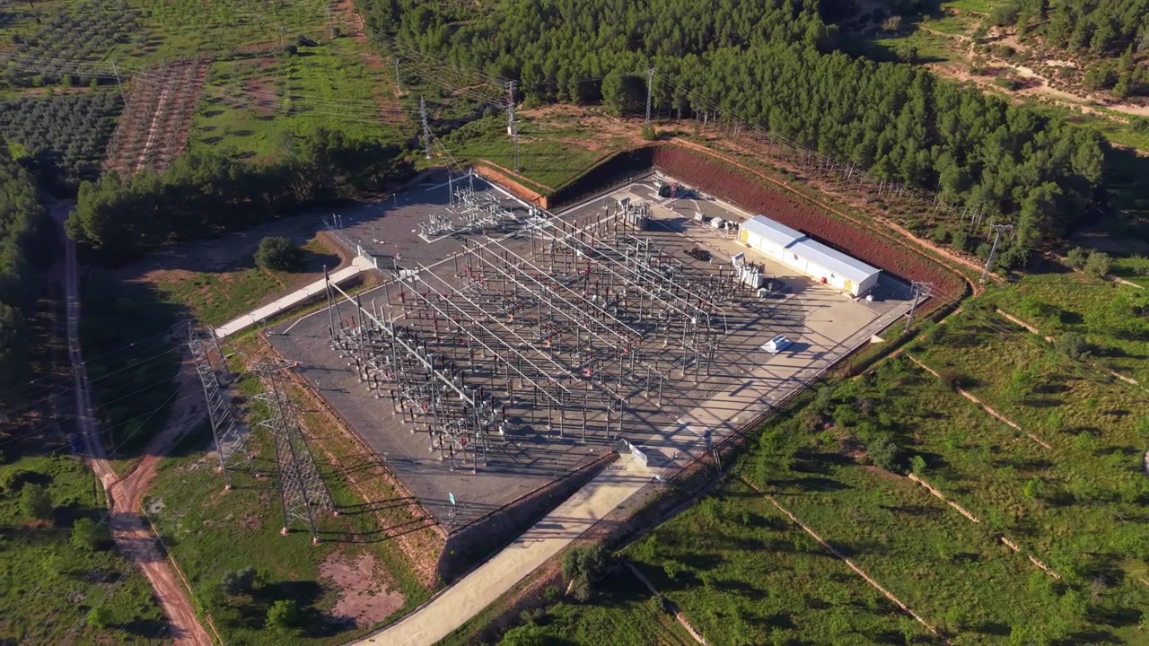 High voltage electrical substation surrounded by green pine forest hills, featuring transmission towers, power lines, transformers and switchgear viewed from above