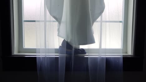 slow slider push in of the bride's wedding veil and dress draped over her shoes resting on a large window sill